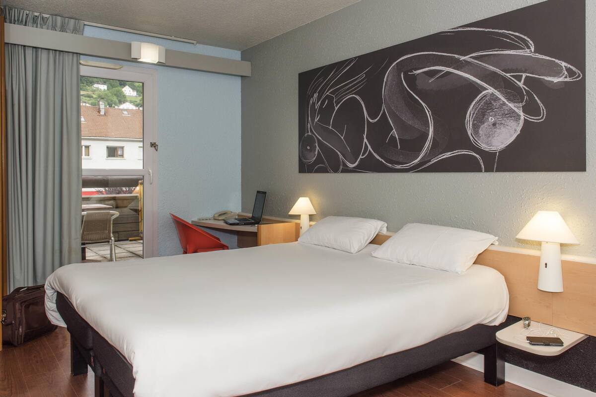 Quarto de hotel com cama, mesas de cabeceira, escrivaninha e uma cadeira, janela e uma obra de arte na parede.