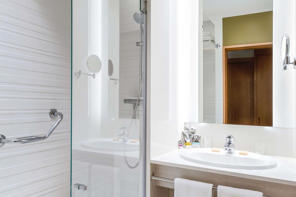 A modern bathroom with a glass-enclosed shower, a white sink with a mirror, and a wooden door.