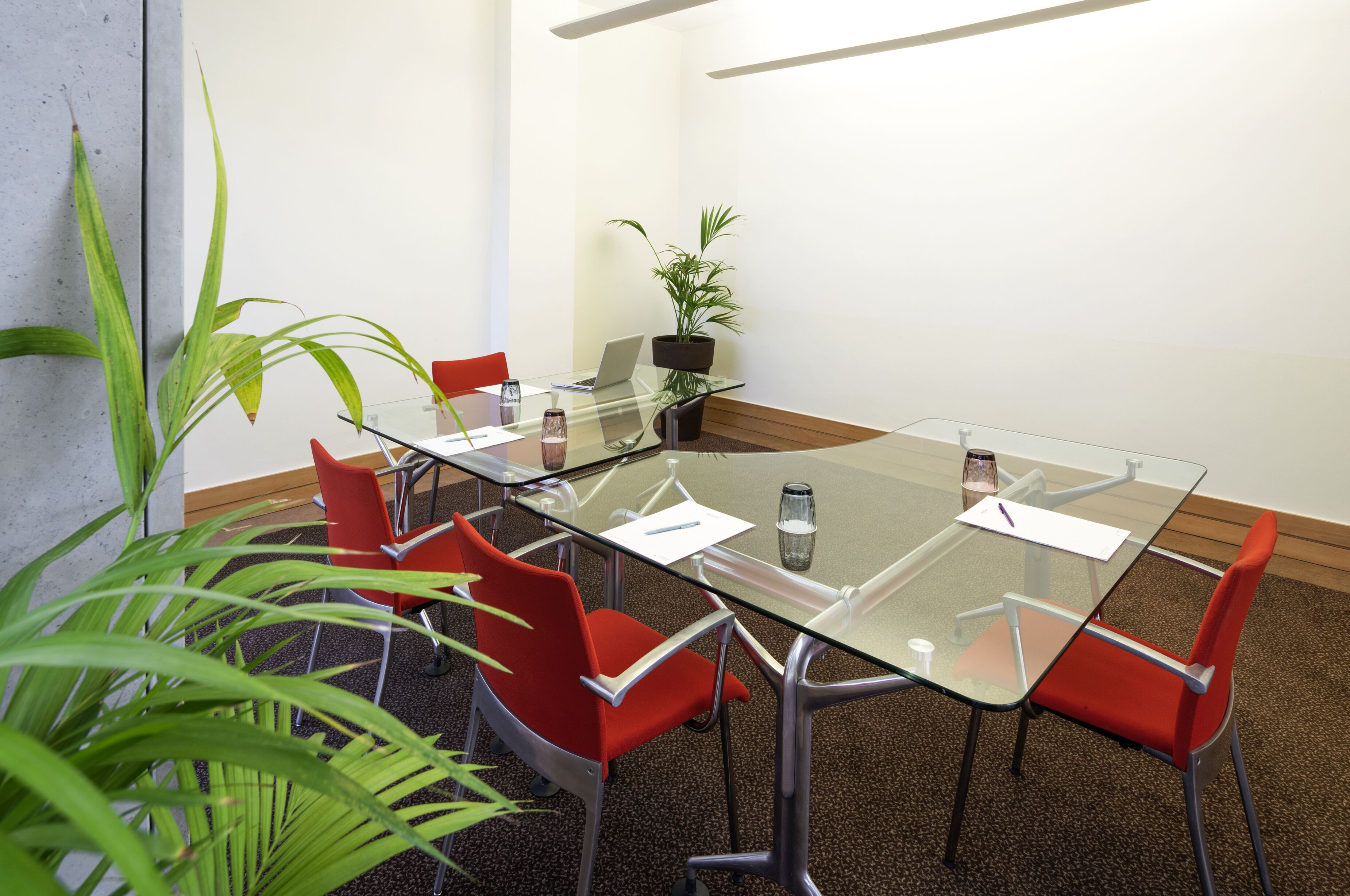 Un salón de conferencias con una mesa de vidrio, sillas rojas, una planta en maceta y un ordenador portátil sobre la mesa.