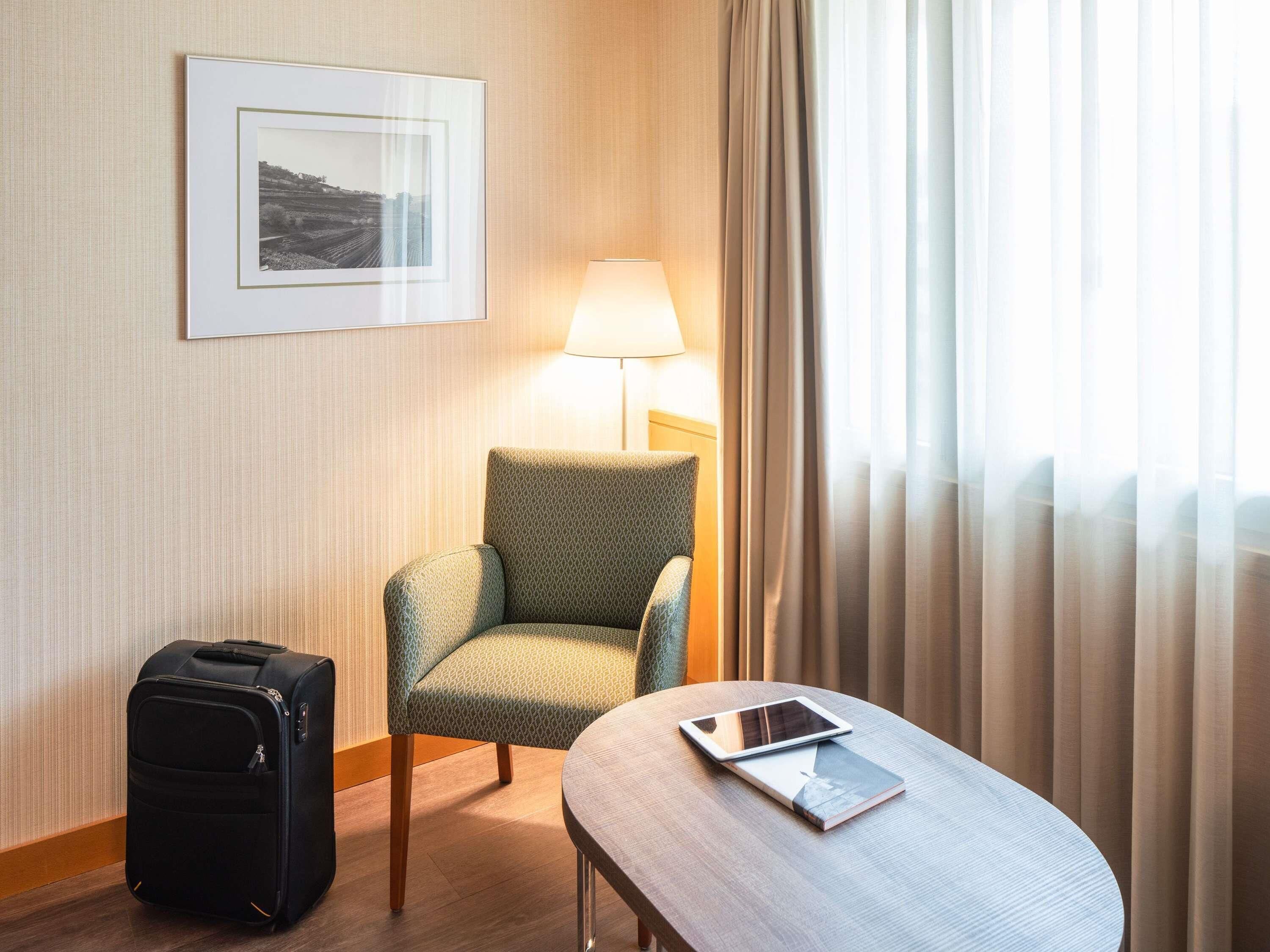 A hotel room with a green patterned armchair, a bedside table with a tablet, a black suitcase, and a framed picture on the wall.