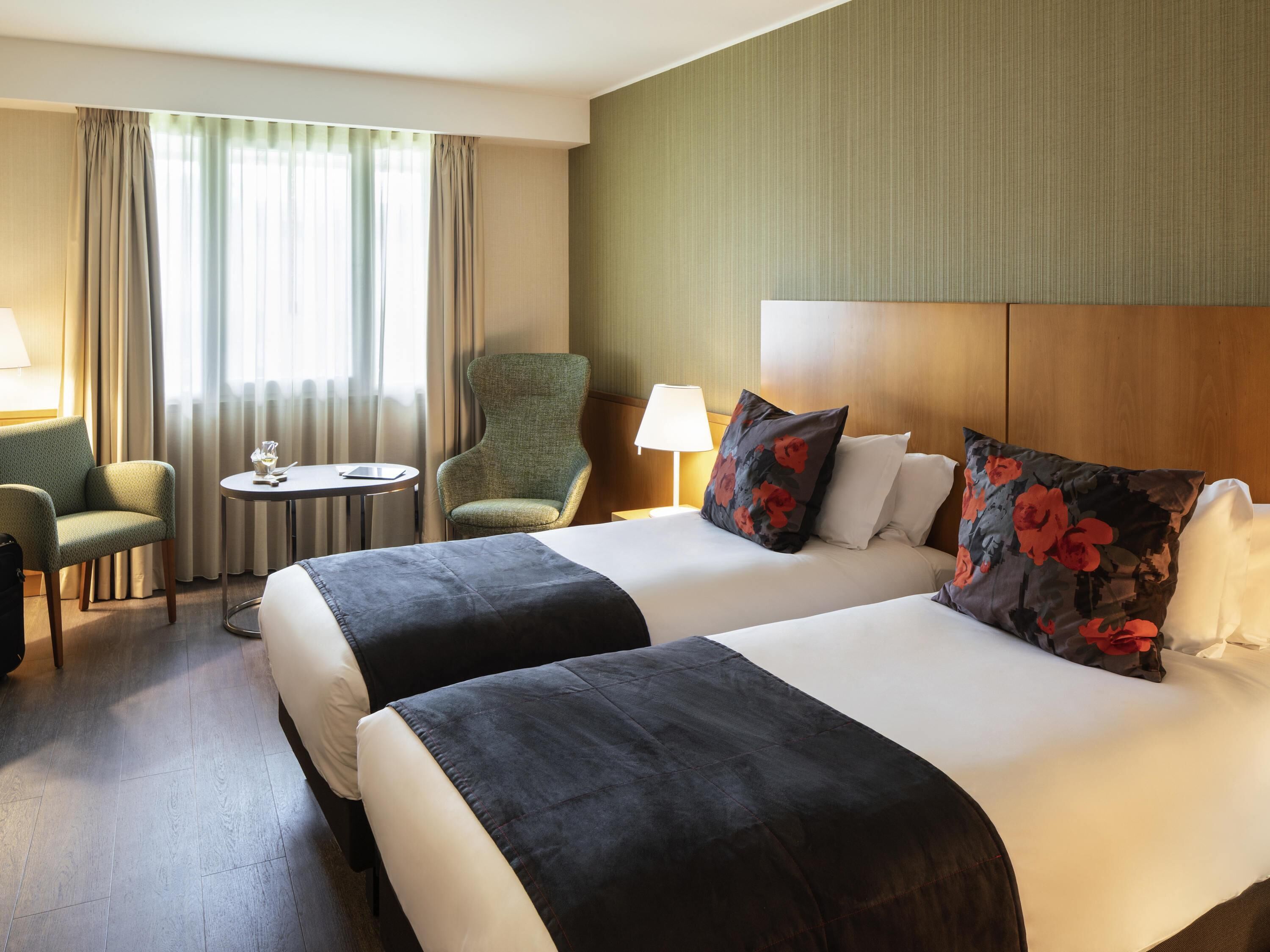 A hotel room with two beds, a chair, a small table, and a lamp.