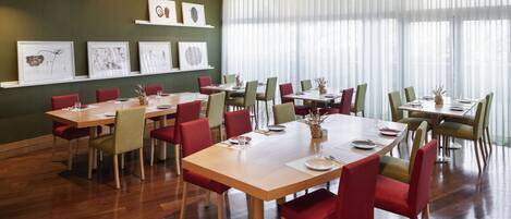 Un comedor con varias mesas y sillas, obras de arte enmarcadas en las paredes y ventanas amplias con cortinas ligeras.