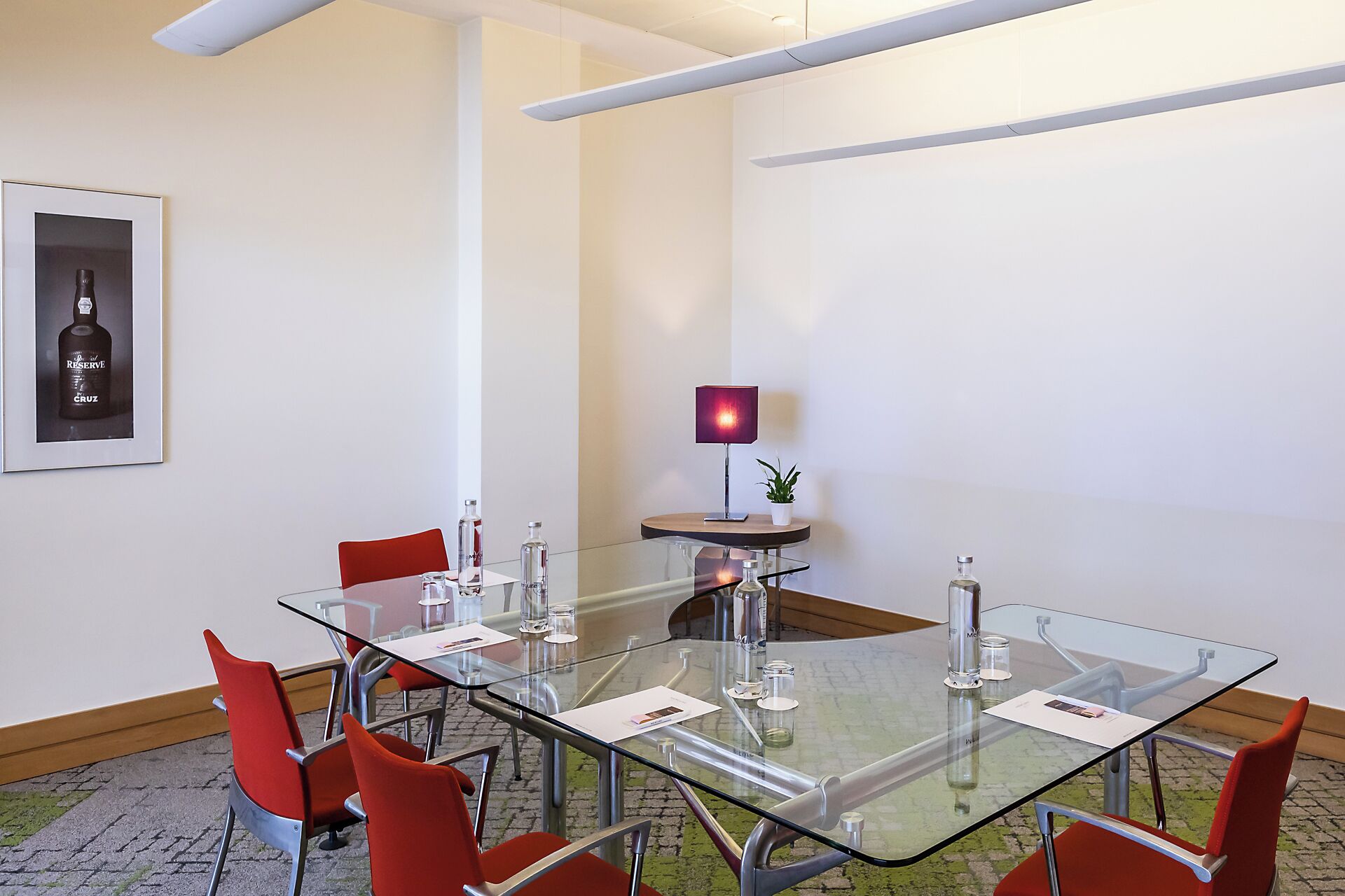 Un salón de conferencias con una mesa de cristal, sillas rojas, una botella de vino enmarcada, una lámpara roja y una pequeña planta en maceta.