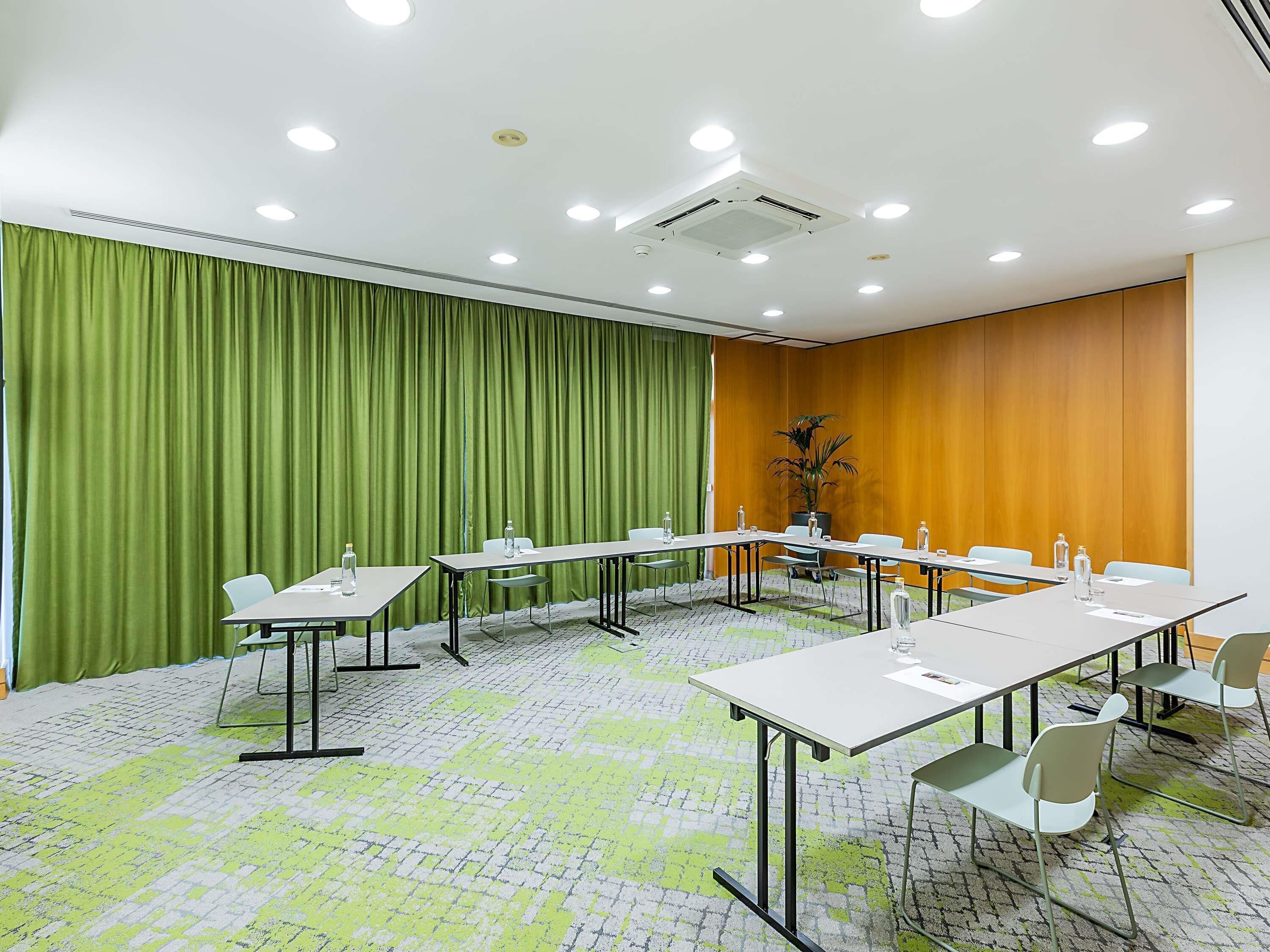 Sala de conferência com uma mesa comprida, cadeiras, um vaso de planta e uma cortina verde.