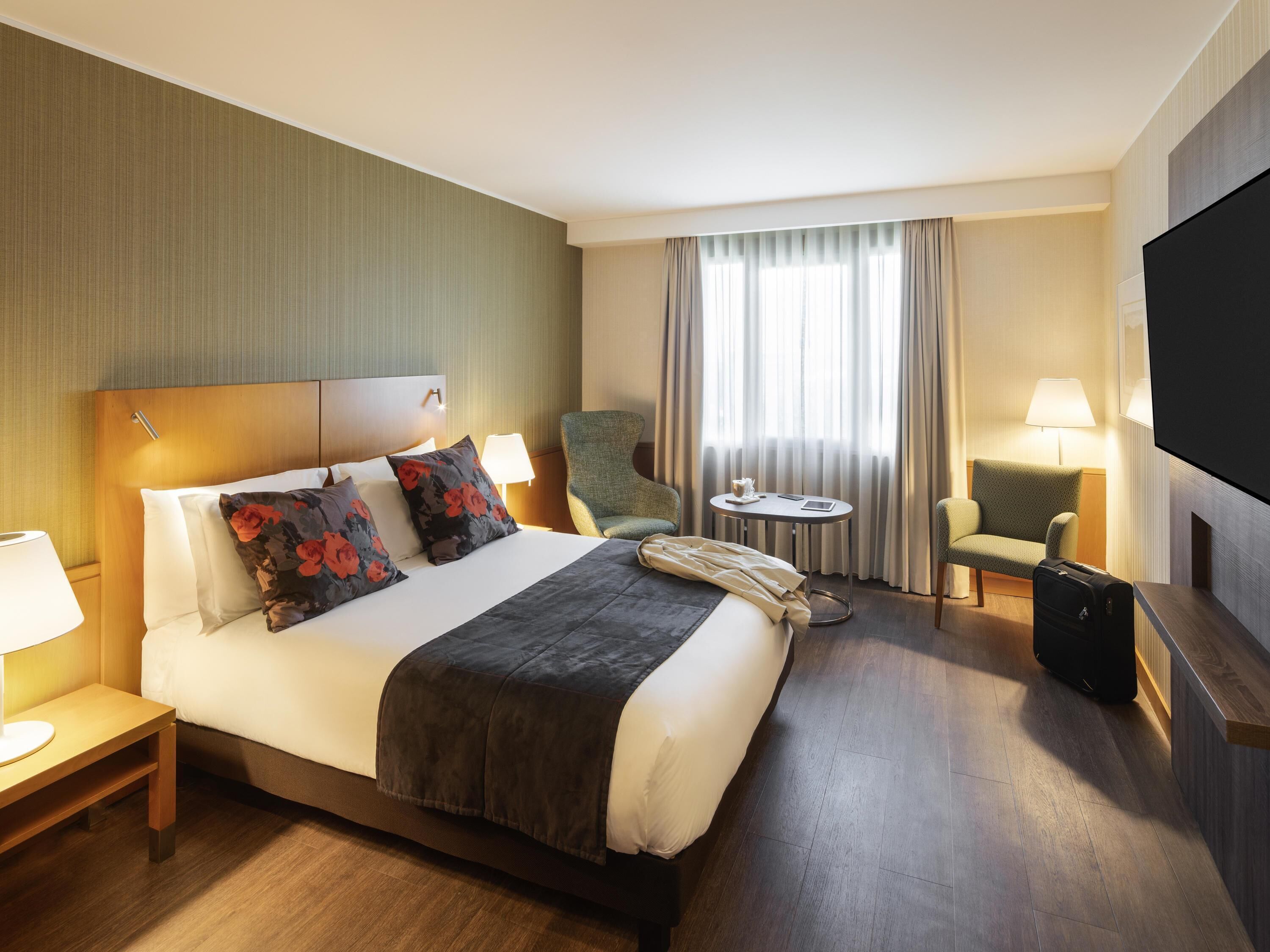 A hotel room with a bed, a chair, a small table, a television, and a suitcase.