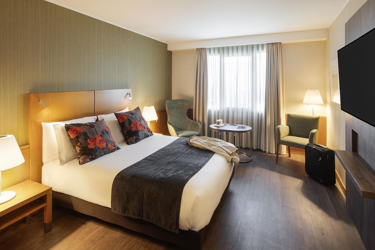 Ein Hotelzimmer mit Bett, Sessel, kleinem Tisch, Fernseher und Koffer.