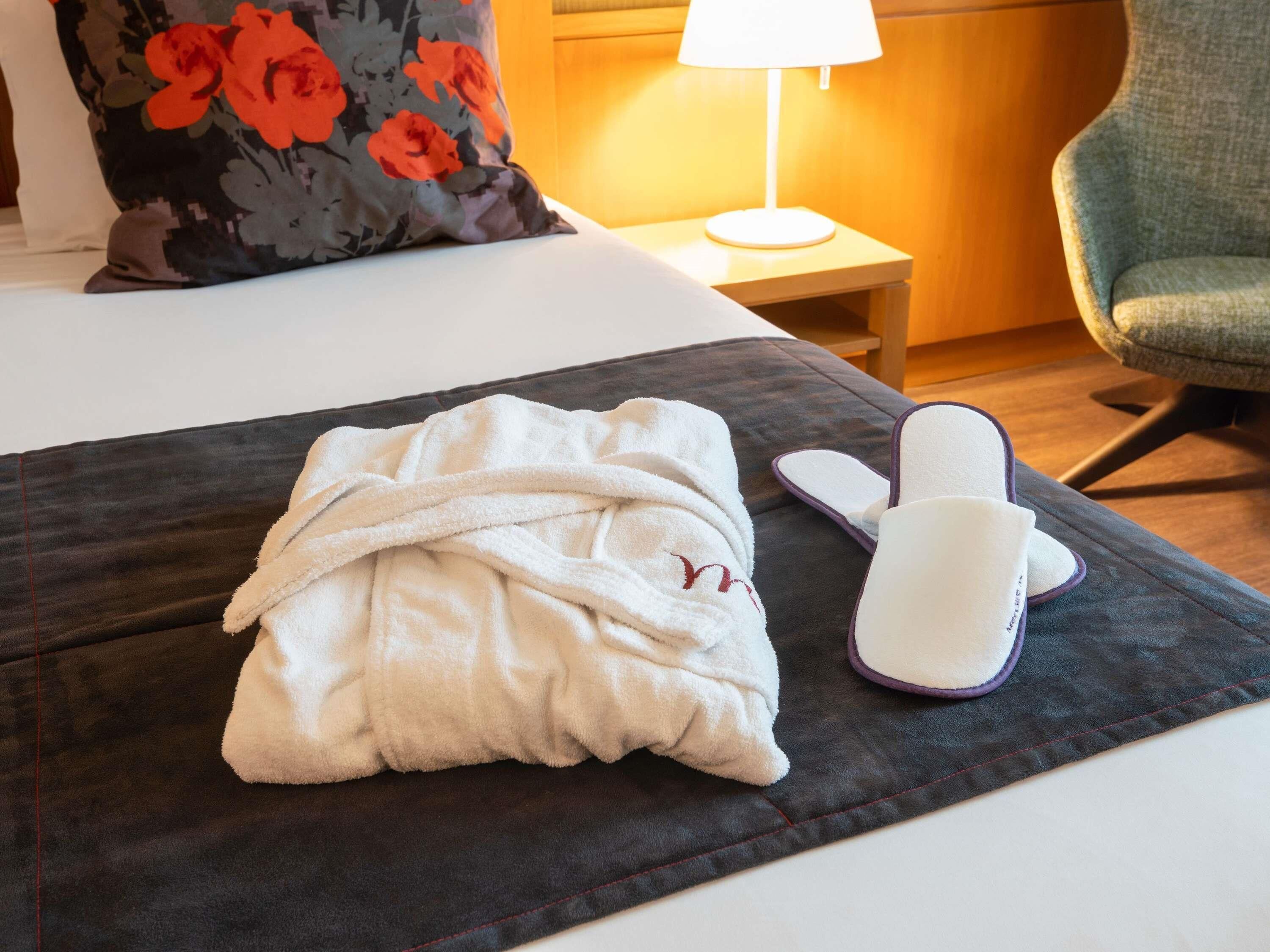 A hotel room with a bed, a pillow, a nightstand with a lamp, a chair, and a robe and slippers on a table.