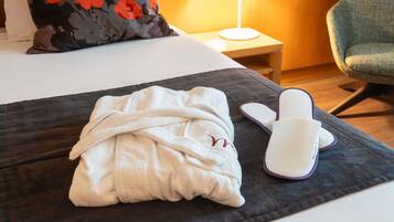 Une chambre d’hôtel avec un lit, un oreiller, une table de chevet avec une lampe, une chaise, et une robe de chambre et des chaussons posés sur une table.