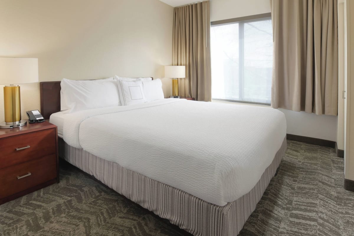 A hotel room with a large bed, two bedside lamps, a wooden nightstand, and a window with curtains.
