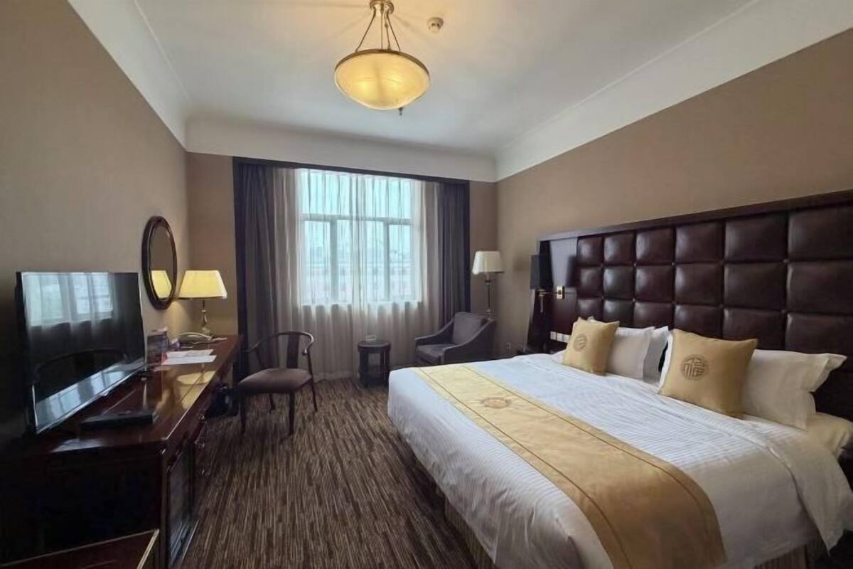 A hotel room with a large bed, a television, a desk with a lamp, a chair, and a window with curtains.