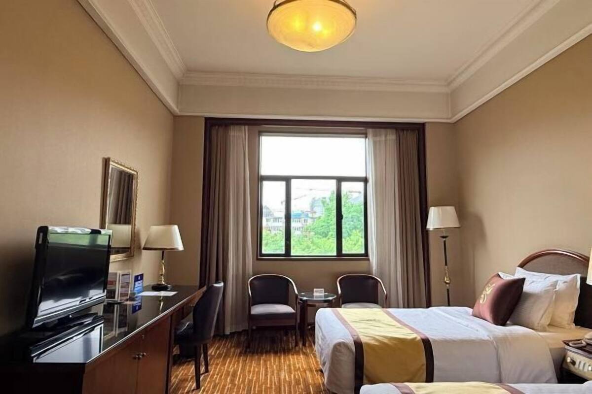 A hotel room with a bed, a desk with a television, a chair, a small table, and a window with curtains.