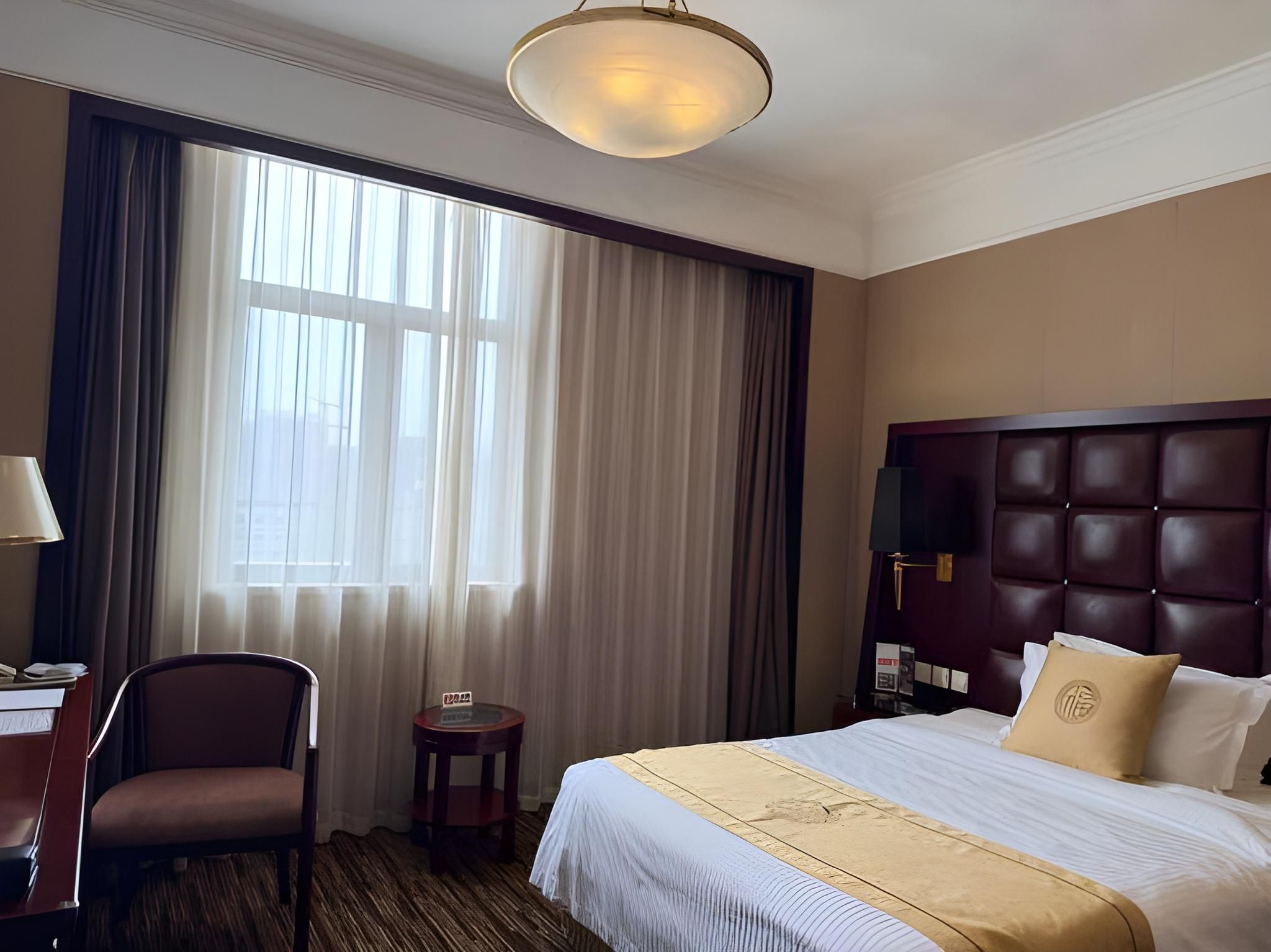 A hotel room with a large bed, a desk with a chair, a small table, and a window with curtains.