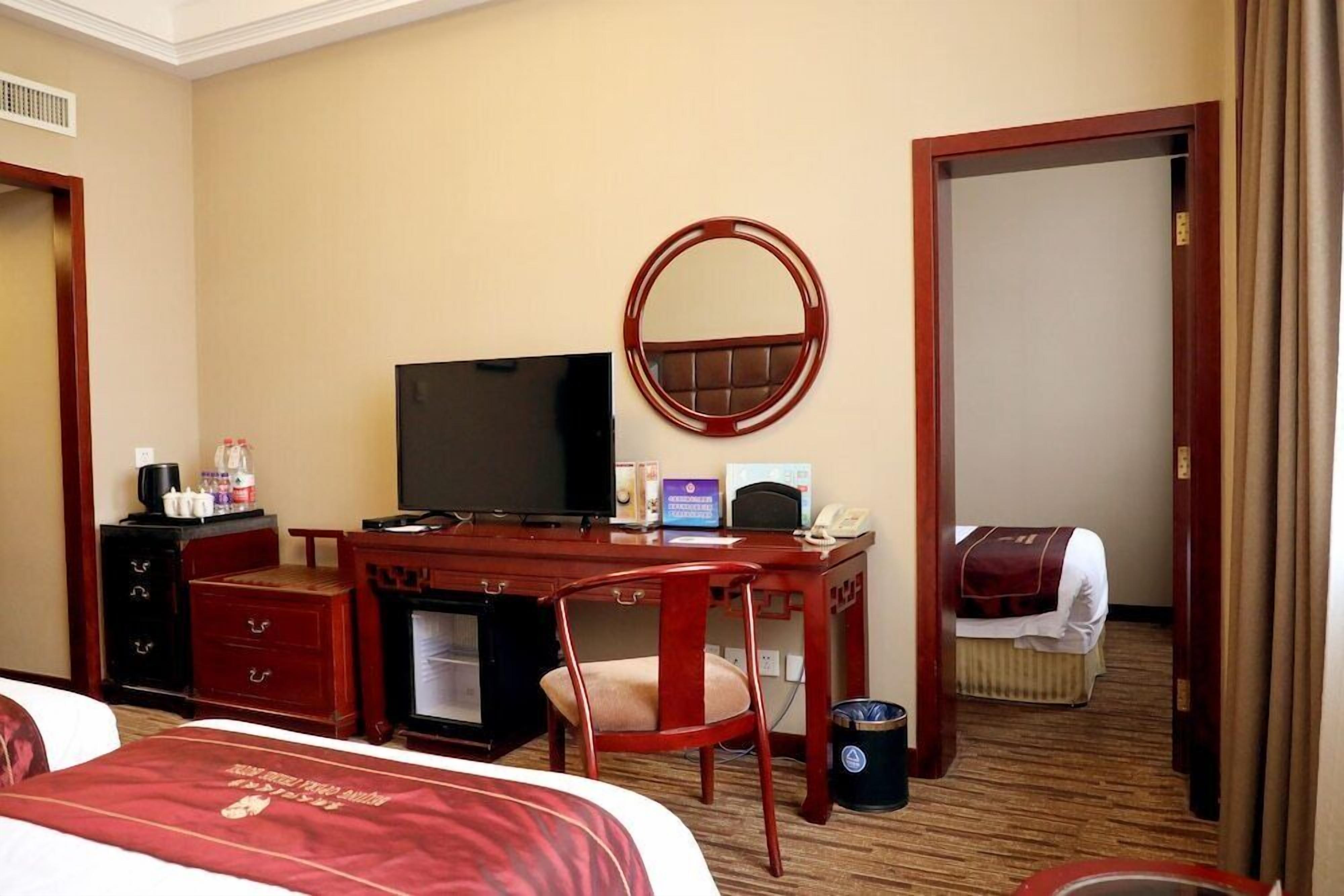 A hotel room with a bed, a desk with a computer, a television, a mirror, a chair, a small table with a telephone, and a waste bin.