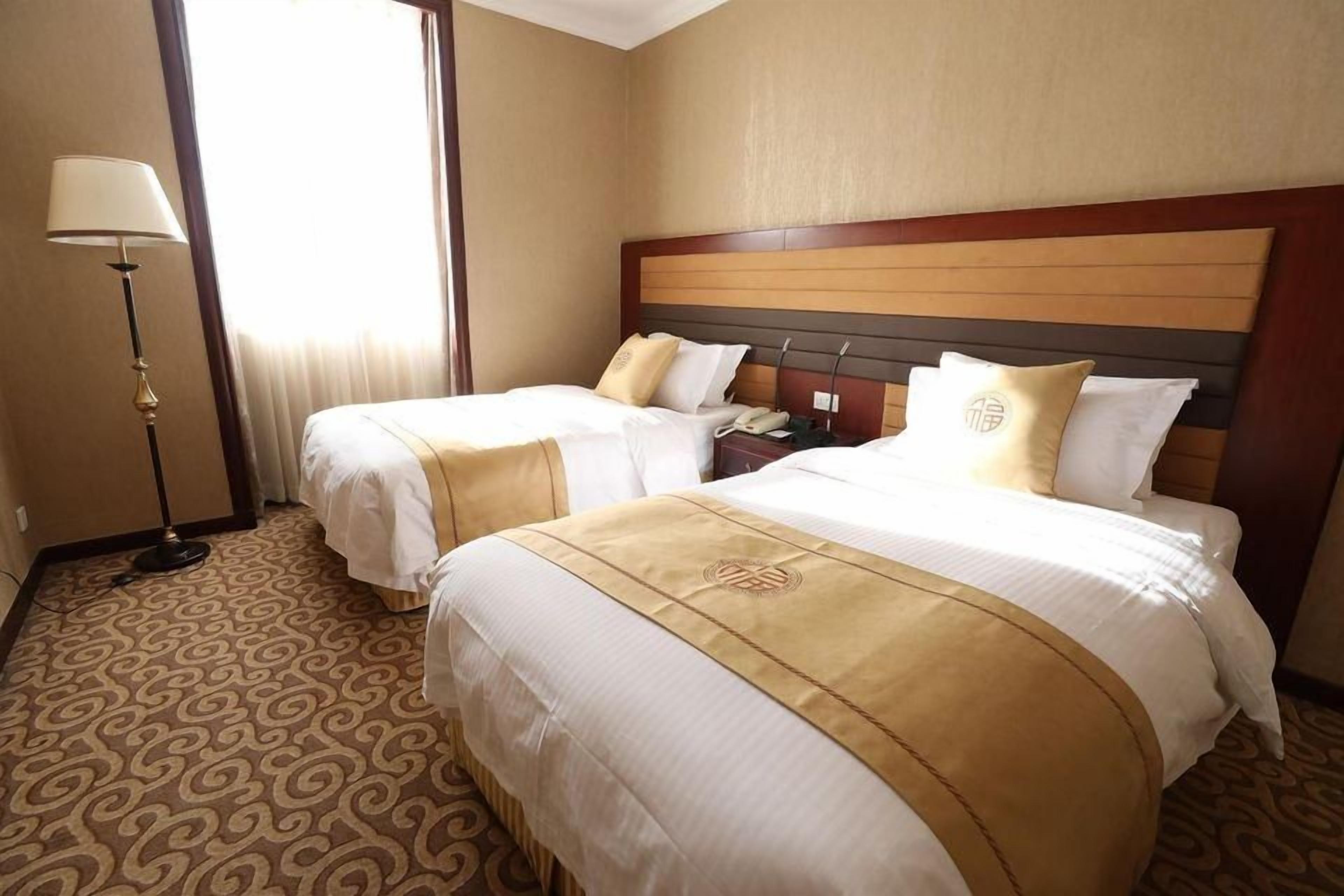 Quarto de hotel com duas camas, mesa de cabeceira, abajur e carpete estampado.