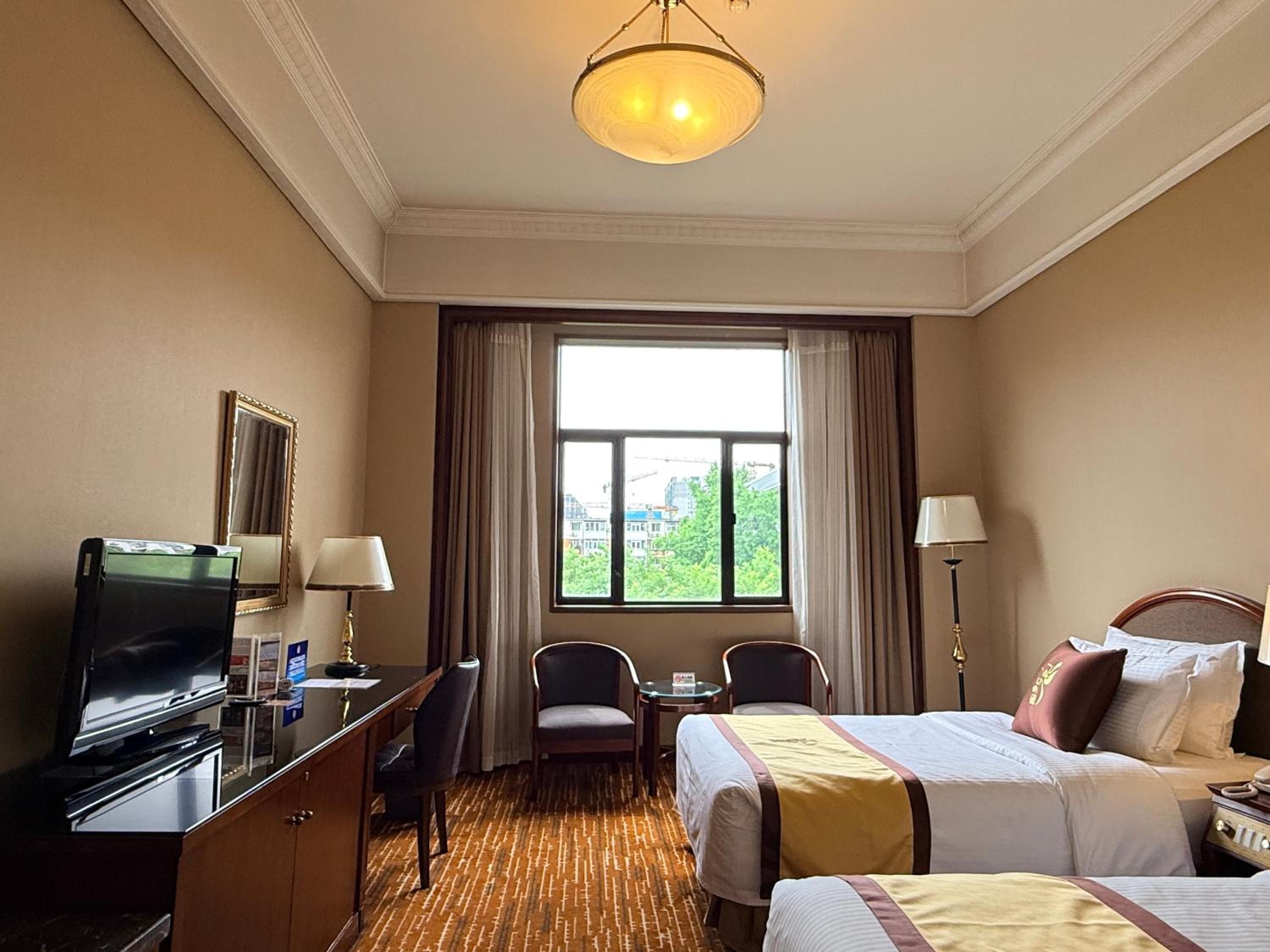 Ein Hotelzimmer mit Bett, Schreibtisch mit Fernseher, Stuhl, Lampe und Blick auf Gebäude durch das Fenster.