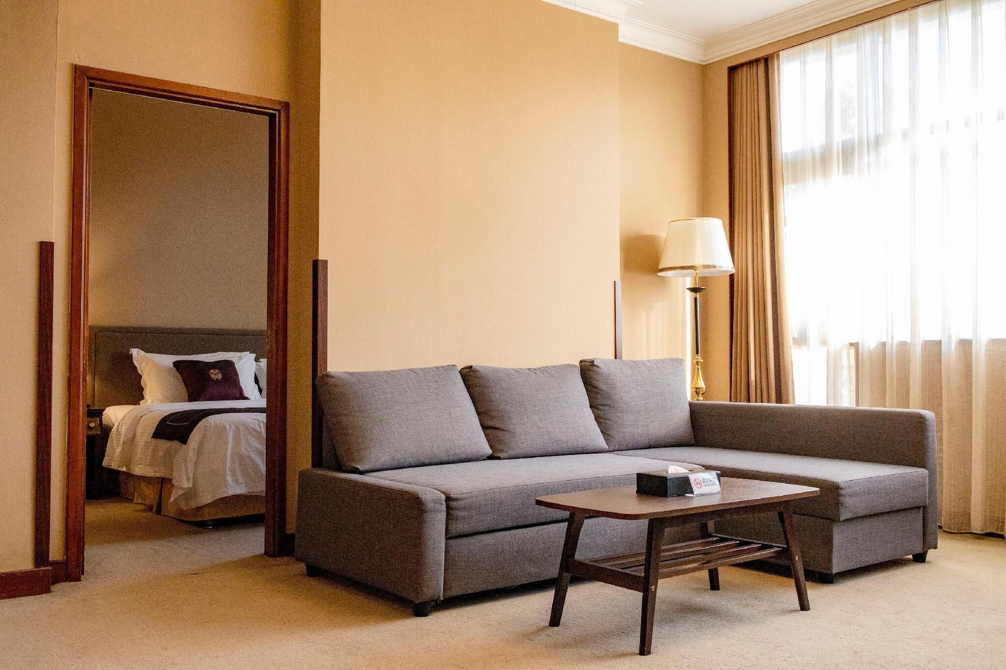 Una habitación de hotel con un sofá gris, una mesita de madera, una lámpara y una cama con ropa de cama blanca y una almohada morada.