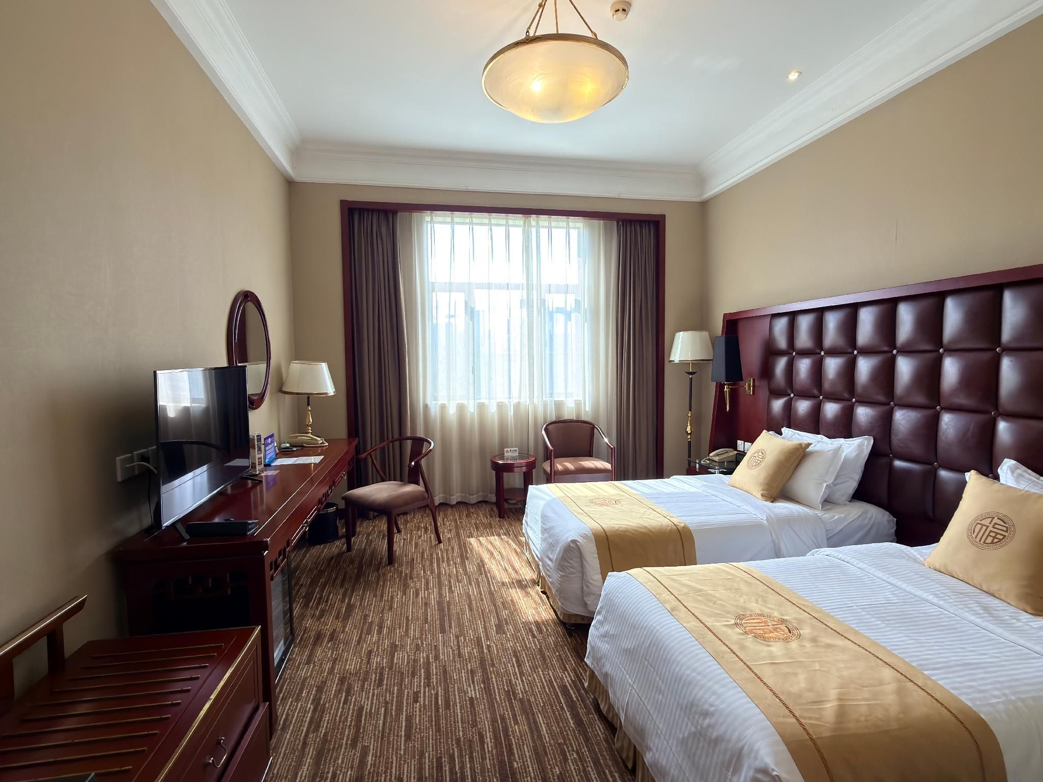 A hotel room with two beds, a desk with a TV, a chair, a small table, and a large window with curtains.