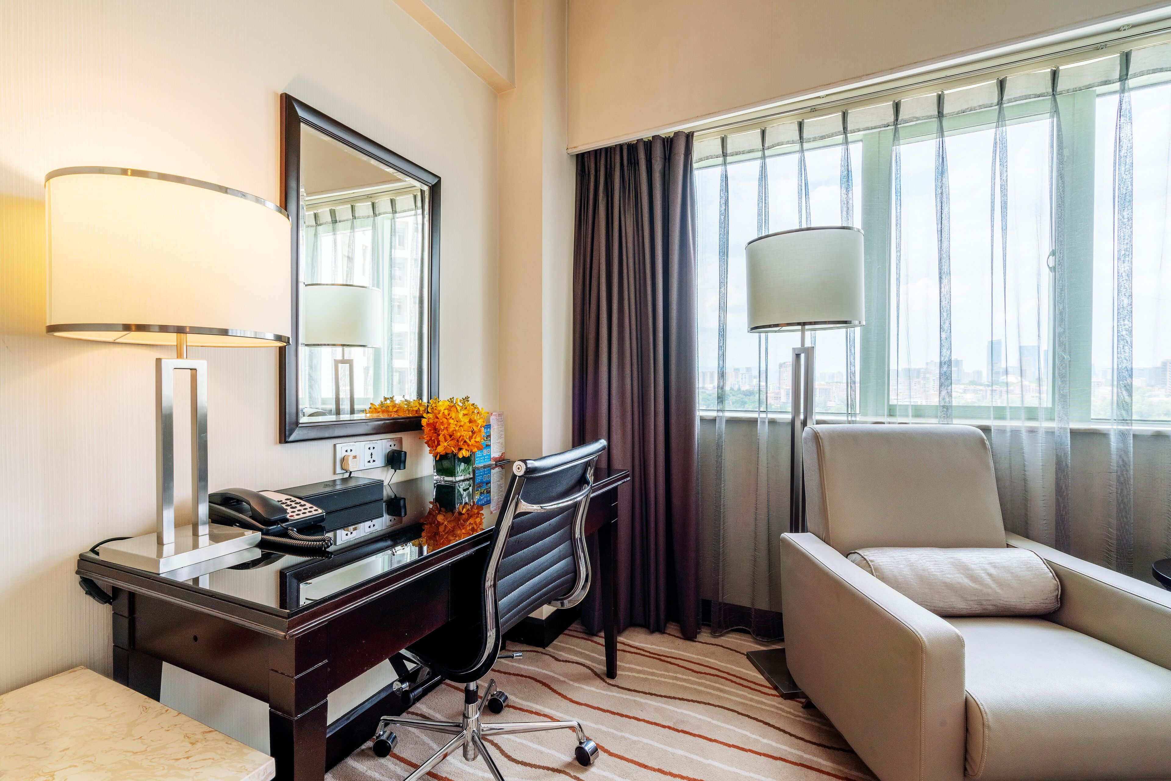 Una habitación de hotel con escritorio, silla, lámpara y un amplio ventanal con cortinas.