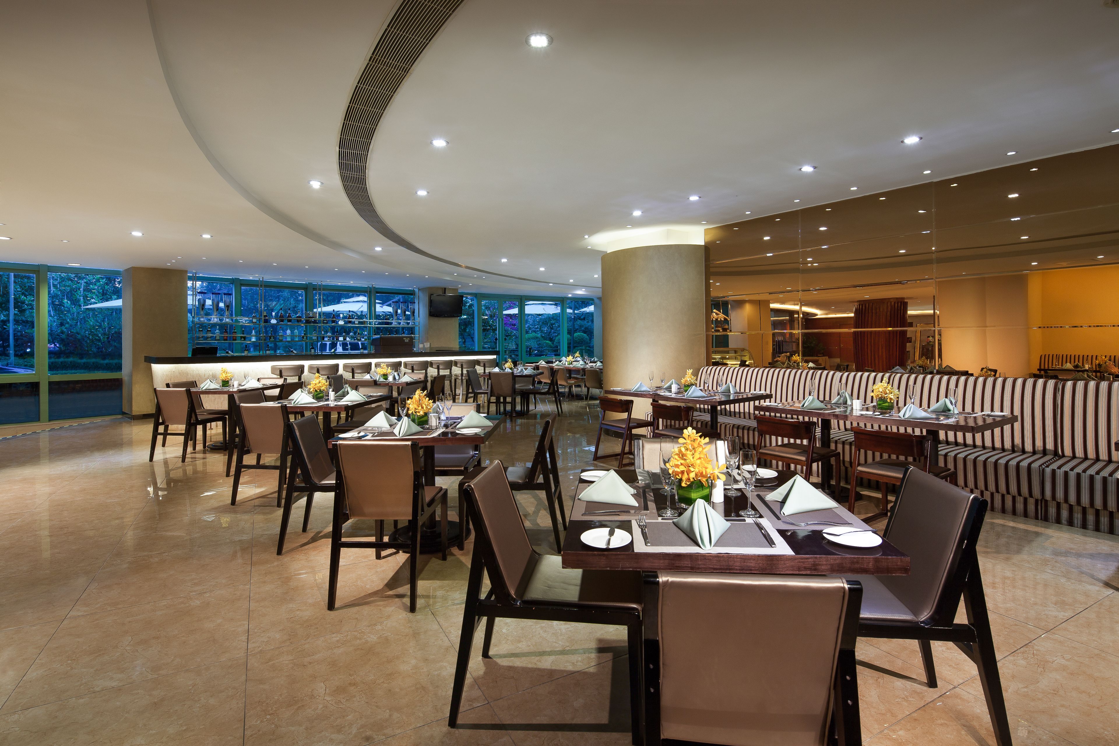 A spacious restaurant with multiple dining tables and chairs, large windows offering a view of the outdoors, and a modern ceiling design.