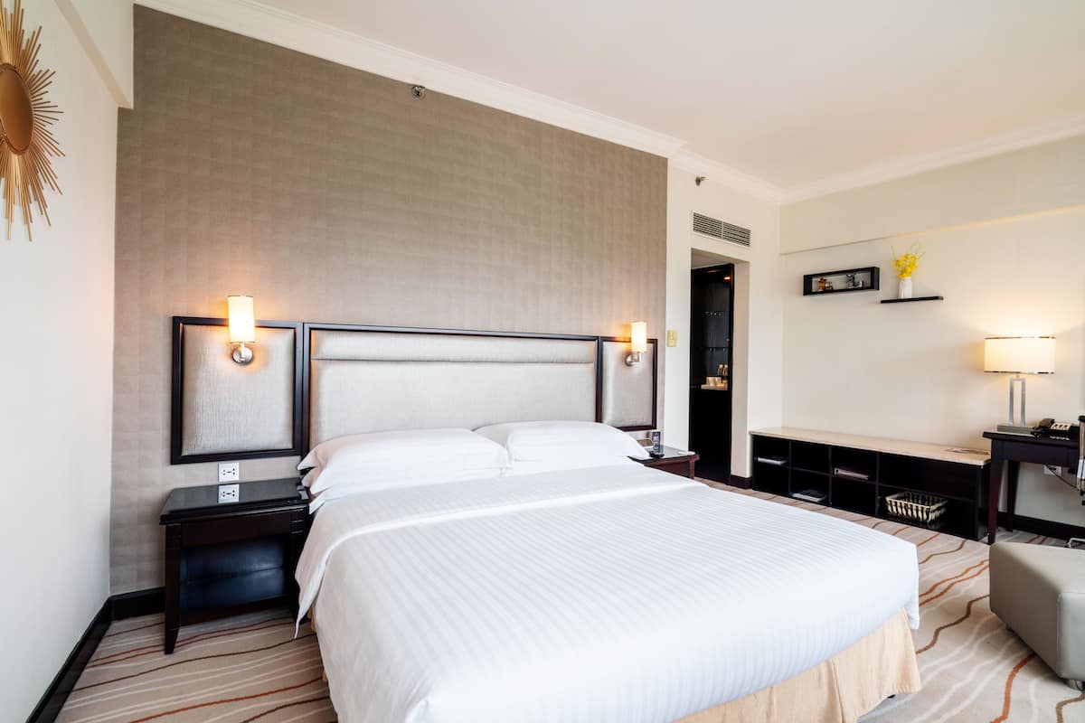 A hotel room with a large bed, two bedside tables with lamps, a desk with a chair, and a small shelf with decorative items.