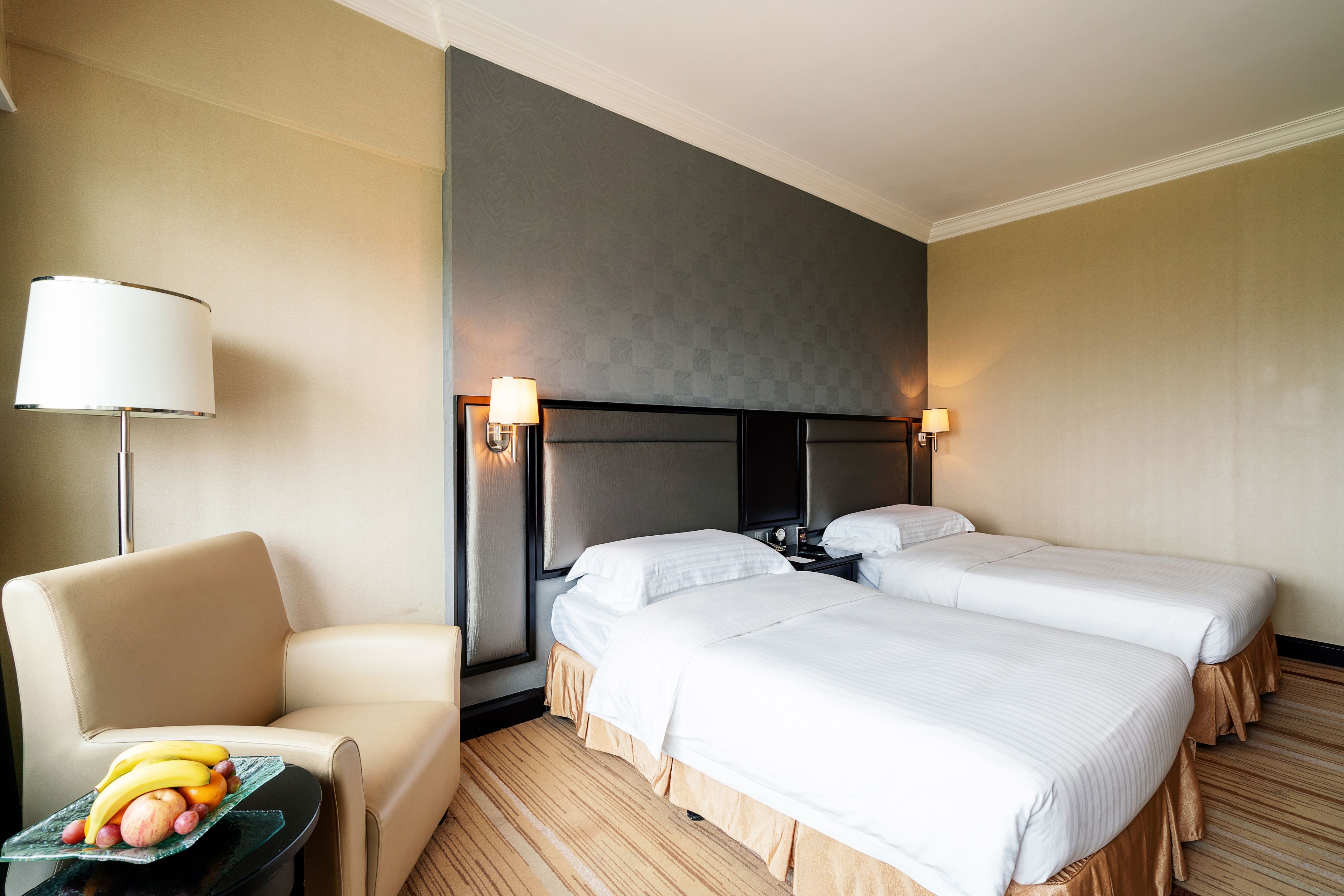 A hotel room with two beds, a chair, a small table with fruit, and a lamp.