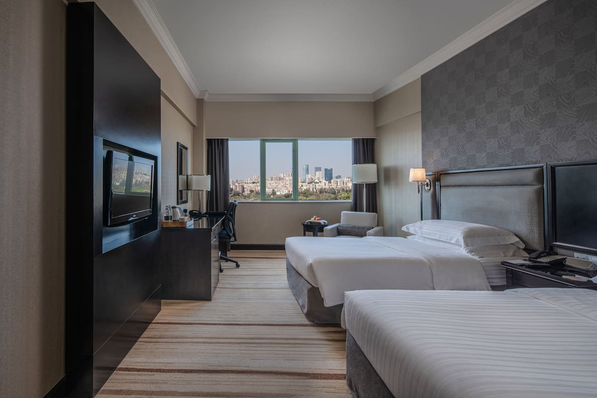 A hotel room with two beds, a desk, a TV, and a city view.