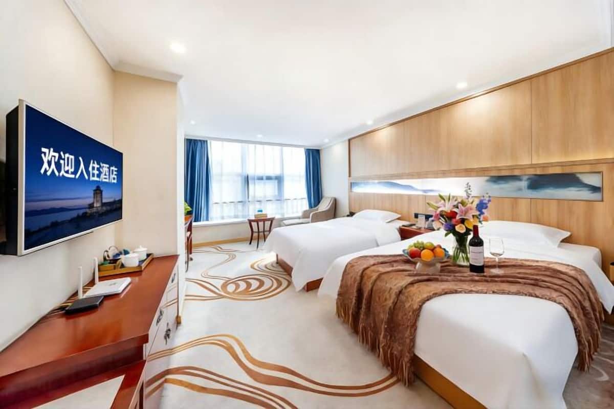 A hotel room with two beds, a TV, a desk, and a large window.