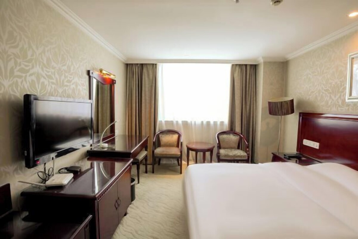 A hotel room with a bed, a desk with a flat-screen TV, two chairs, a small table, and a window with curtains.