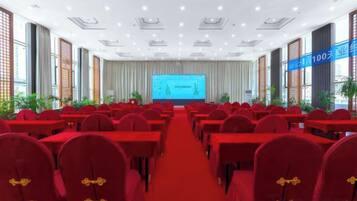 Une salle de conférence avec des rangées de tables et de chaises, un grand écran à l’avant et un tapis rouge.
