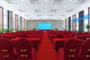 Un salón de conferencias con mesas y sillas dispuestas en filas, una pantalla grande al frente y una alfombra roja.