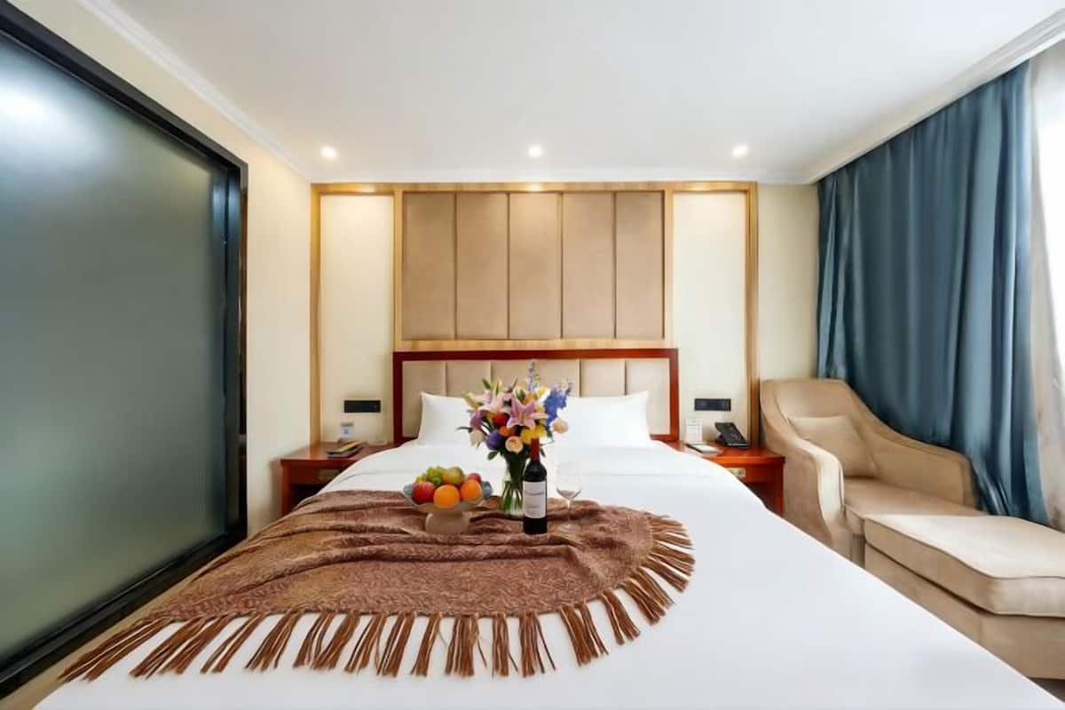 A hotel room with a bed, a bedside table with a vase of flowers, a fruit bowl, and a bottle of wine.
