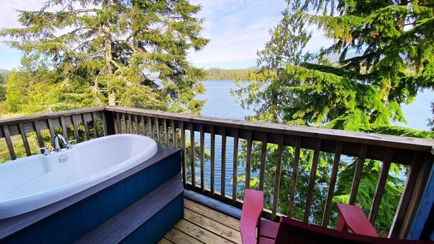 A wooden deck with a hot tub and two chairs overlooking a lake. - Waters Edge Shoreside Suites (Ucluelet)