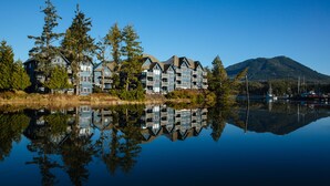 A multi-story hotel with a reflection in the water, surrounded by trees and a mountain in the background. - Waters Edge Shoreside Suites (Ucluelet)