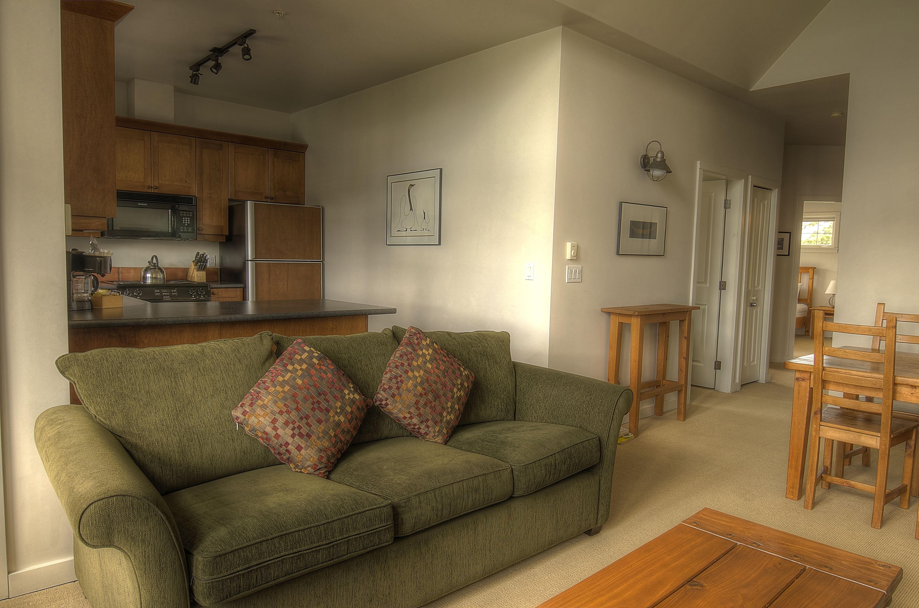 A cozy living room with a green sofa, wooden dining table, and a kitchen in the background.