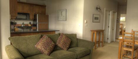 A cozy living room with a green sofa, wooden dining table, and a kitchen in the background.