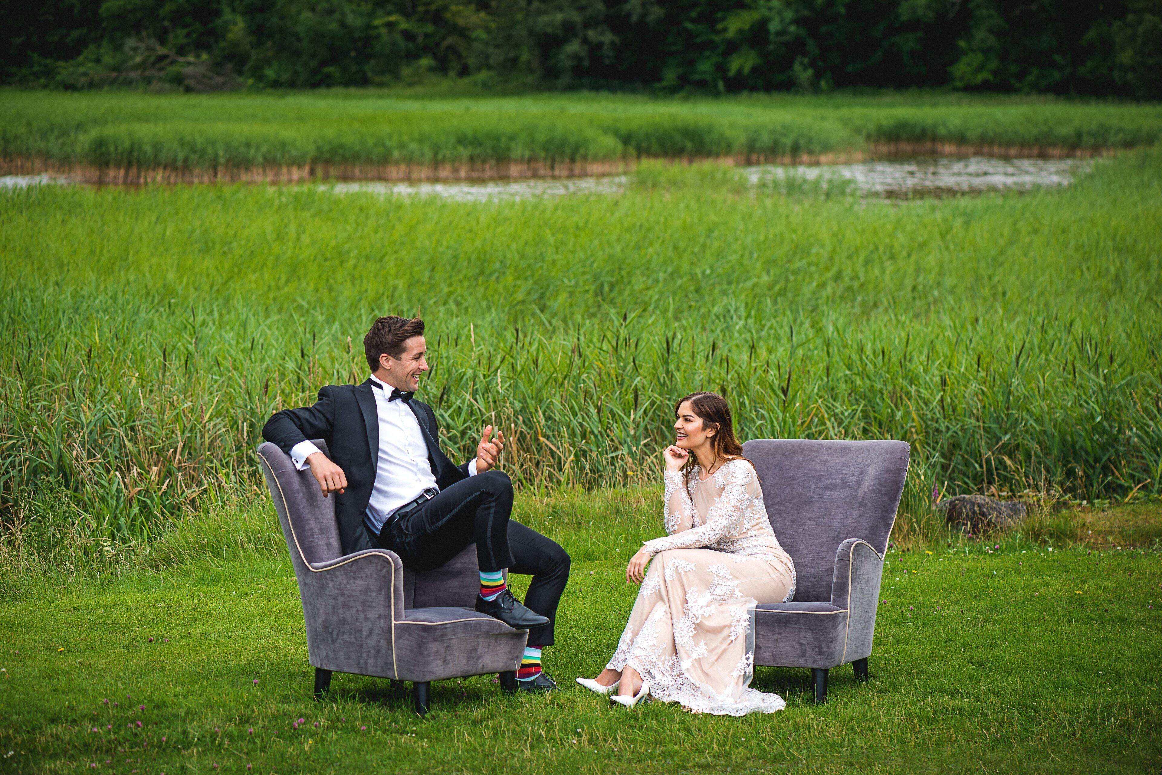 Two people seated on a grey sofa in a grassy field with a pond in the background.