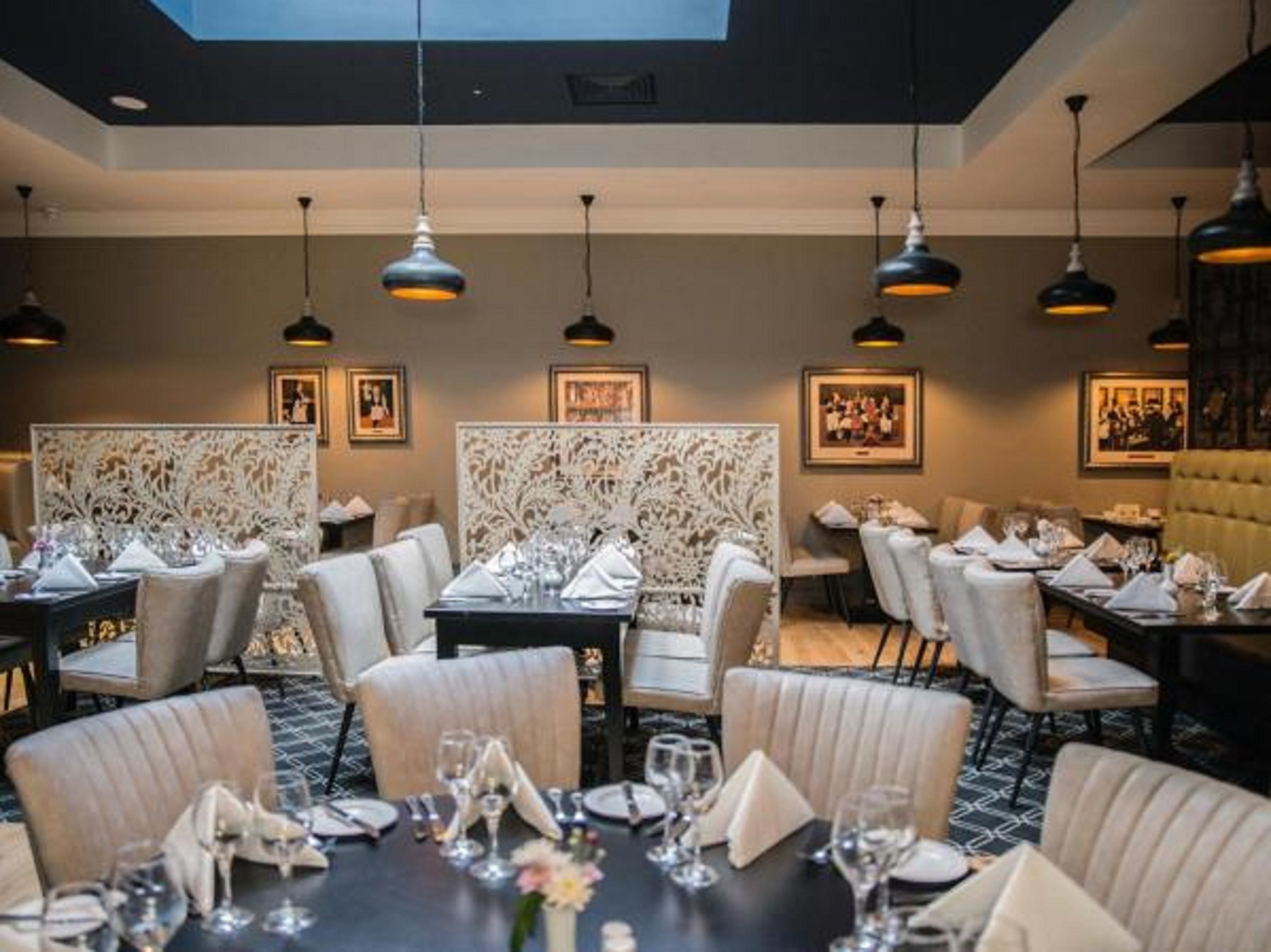 A restaurant with multiple dining tables set with white tablecloths and chairs, overhead pendant lighting, and framed artwork on the walls.