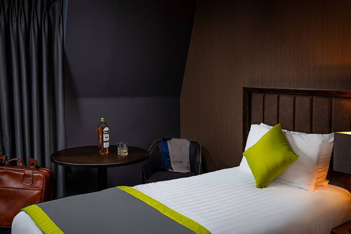 Een hotelkamer met een bed, een kleine tafel met een fles en glas, een stoel en een koffer.