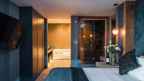 Une chambre d’hôtel moderne avec un grand lit, une salle de bain dotée d’une baignoire et d’une douche. On y trouve une petite table avec un vase de fleurs et une bouteille de vin.