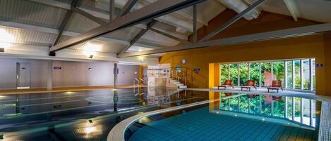 Una piscina interior con suelo de baldosas, una escalera que sube a un nivel superior y vistas a zonas verdes a través de amplias ventanas.