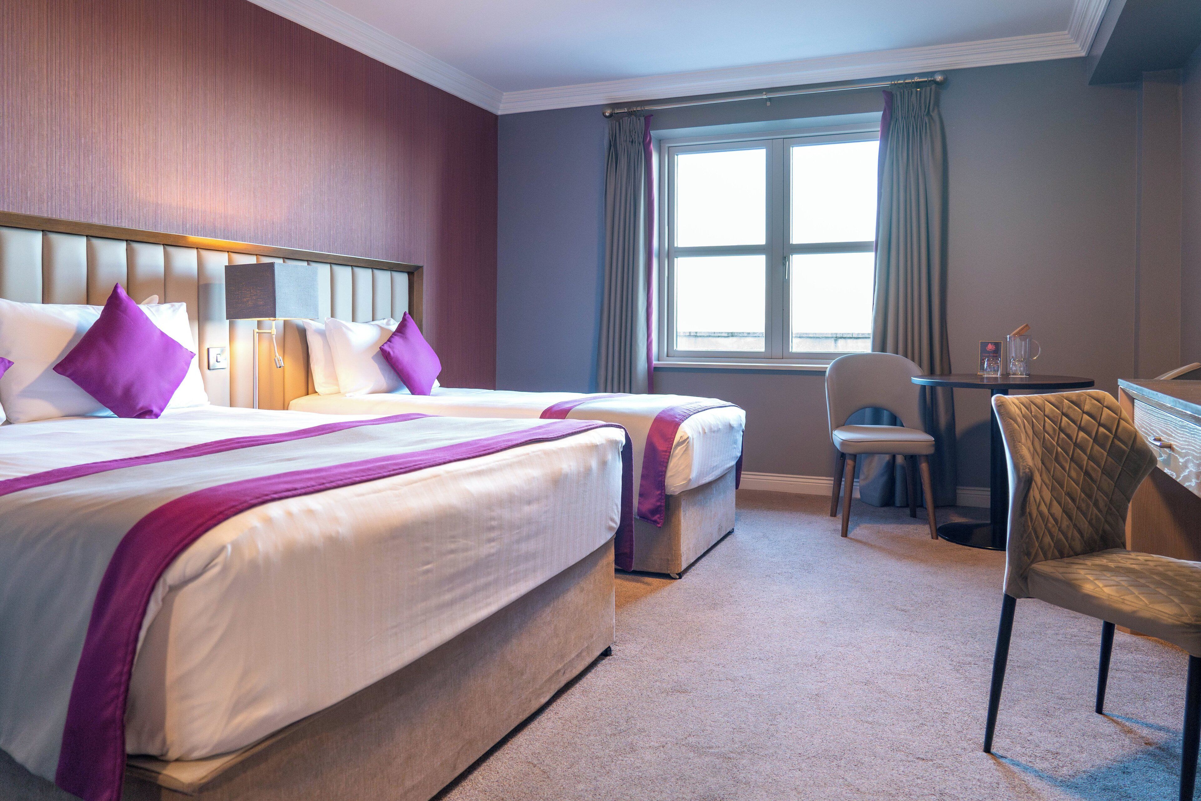 Quarto de hotel com duas camas, uma escrivaninha, uma cadeira e uma janela com cortinas.