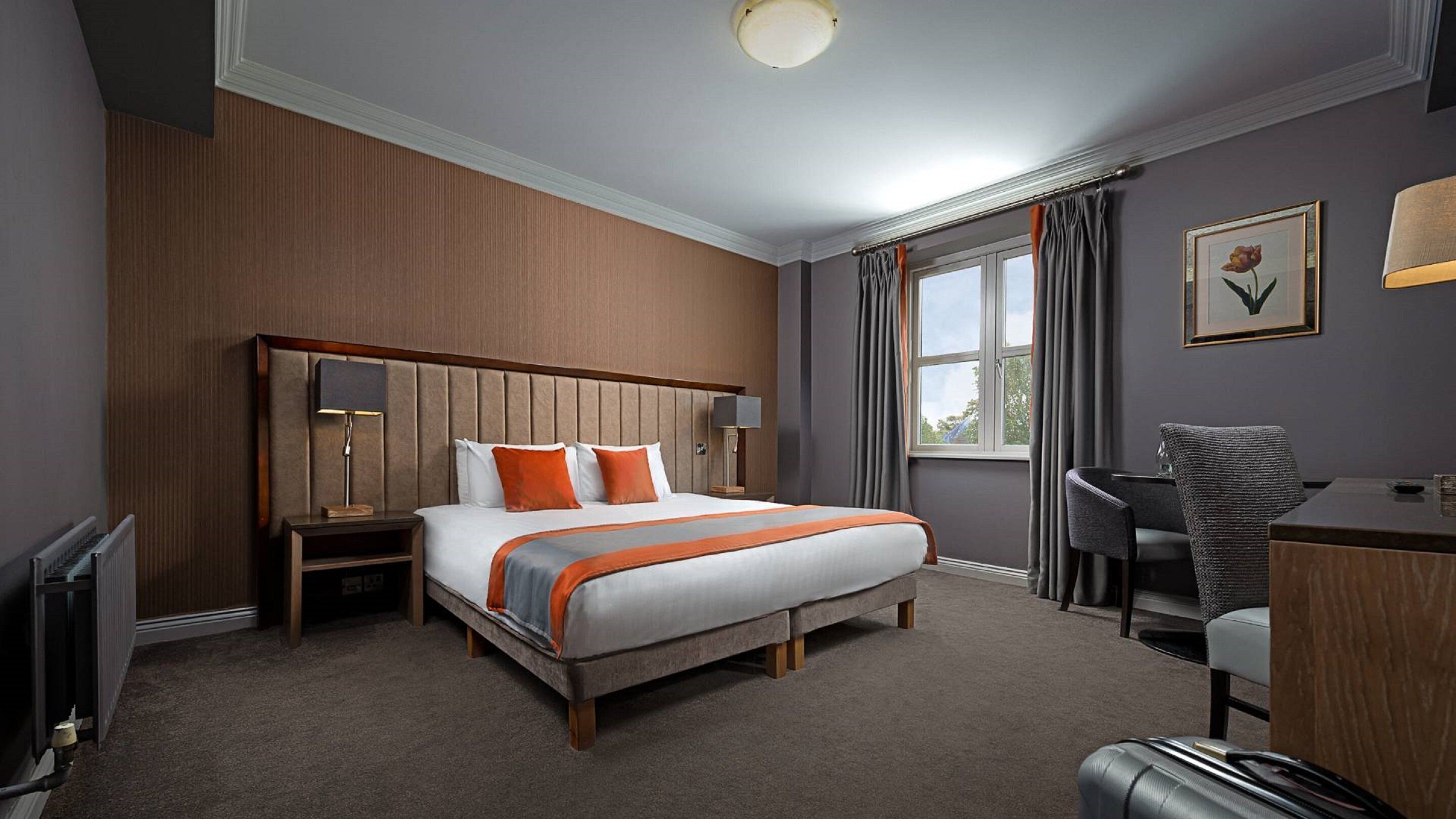 Quarto de hotel com uma cama grande, uma escrivaninha, uma cadeira, um radiador e uma janela com cortinas.