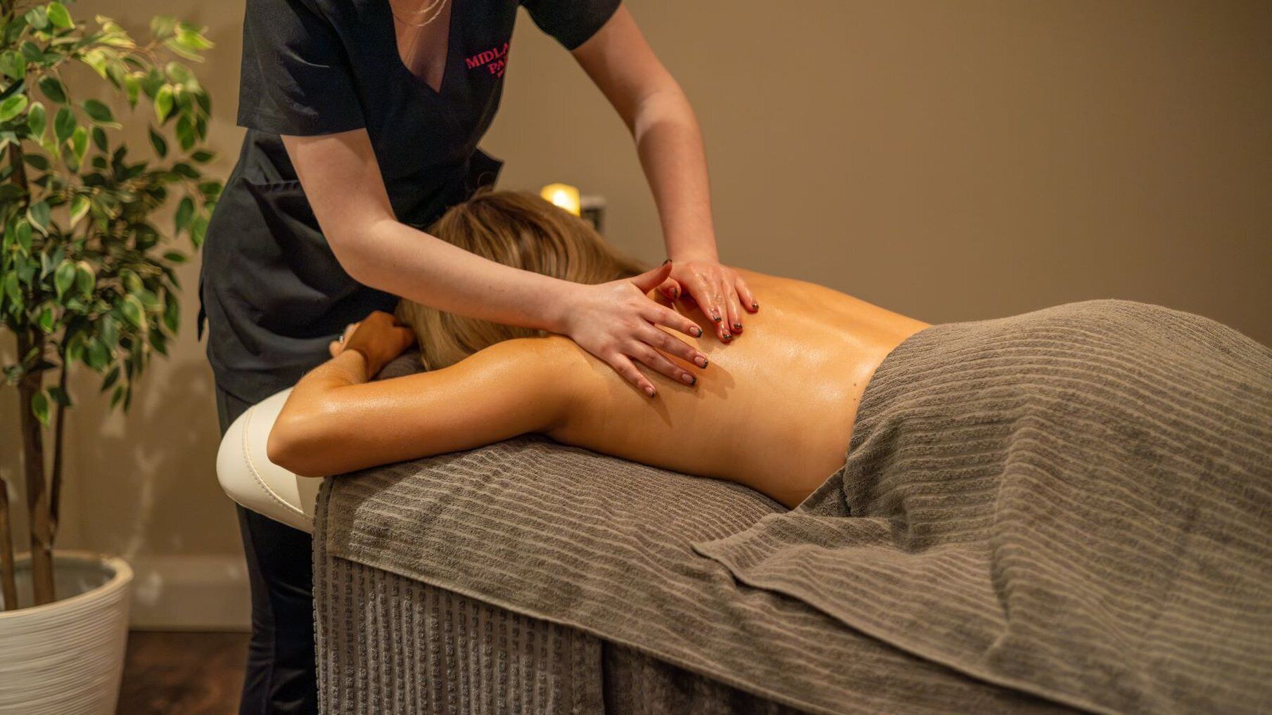 A massage therapy session in a spa setting with a client lying on a treatment table, a therapist attending to the client, and a potted plant in the background.
