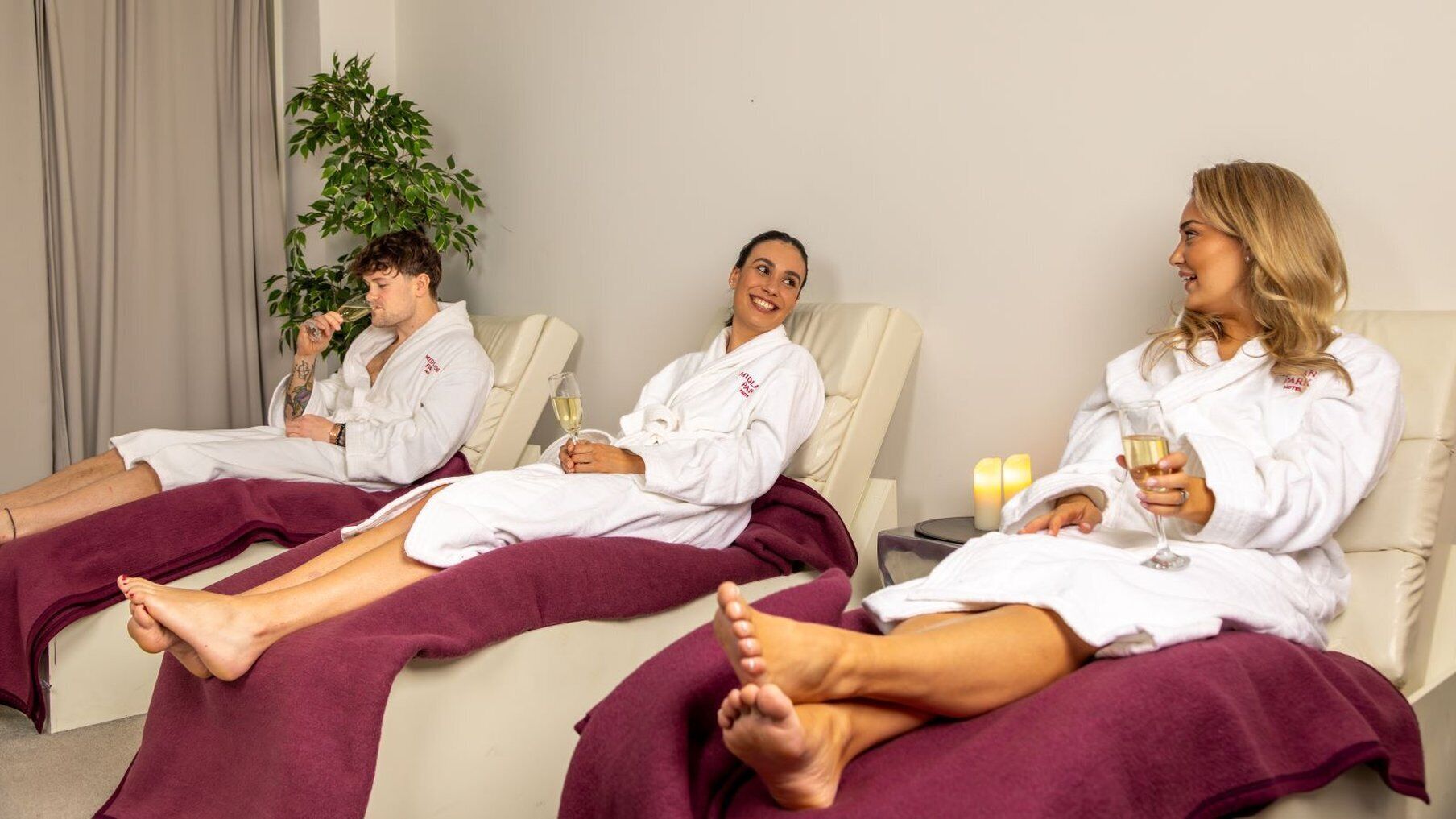 Three people in white robes sitting on separate beds in a room with a plant and candles.