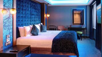 Una habitación de hotel moderna con una cama grande, una mesita de noche con control remoto, un taburete pequeño, un espejo y una pared con un diseño decorativo.