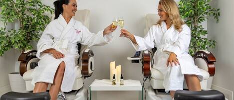 Two individuals in white robes seated in a spa-like setting with foot massage chairs, a small table with candles, and potted plants.