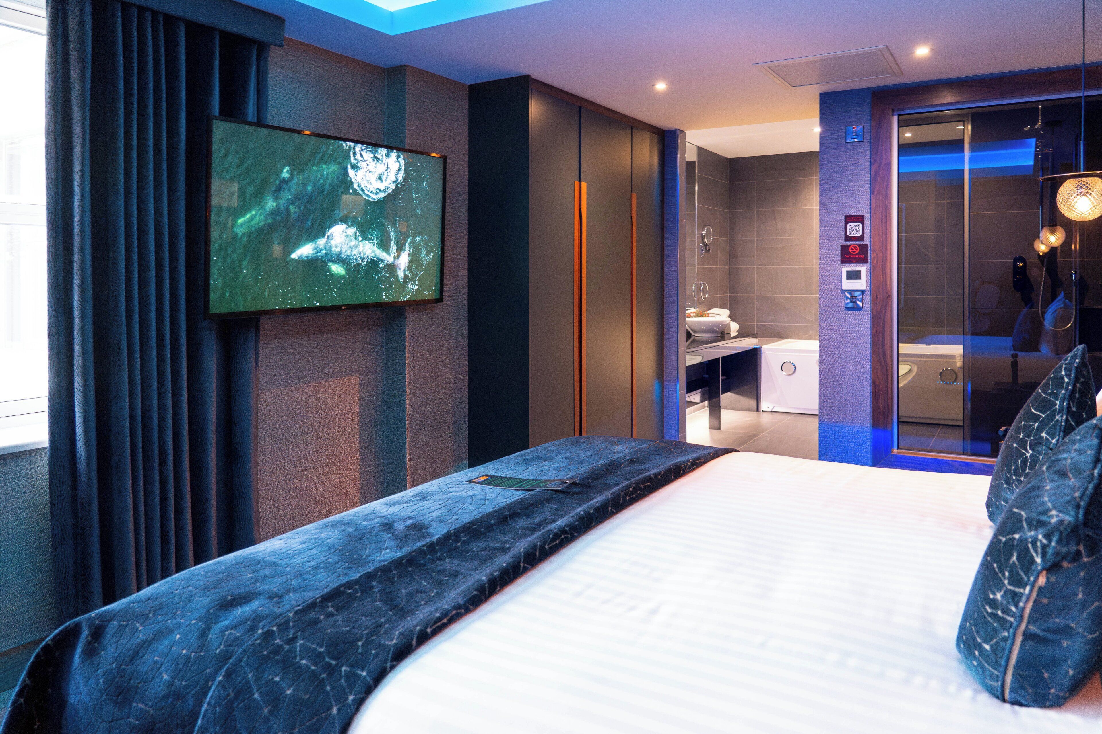 Una habitación de hotel moderna con una cama grande, un televisor montado en la pared, un baño con ducha de vidrio y un panel de control en la pared.