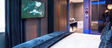 Una habitación de hotel moderna con una cama grande, un televisor montado en la pared, un baño con ducha de vidrio y un panel de control en la pared.