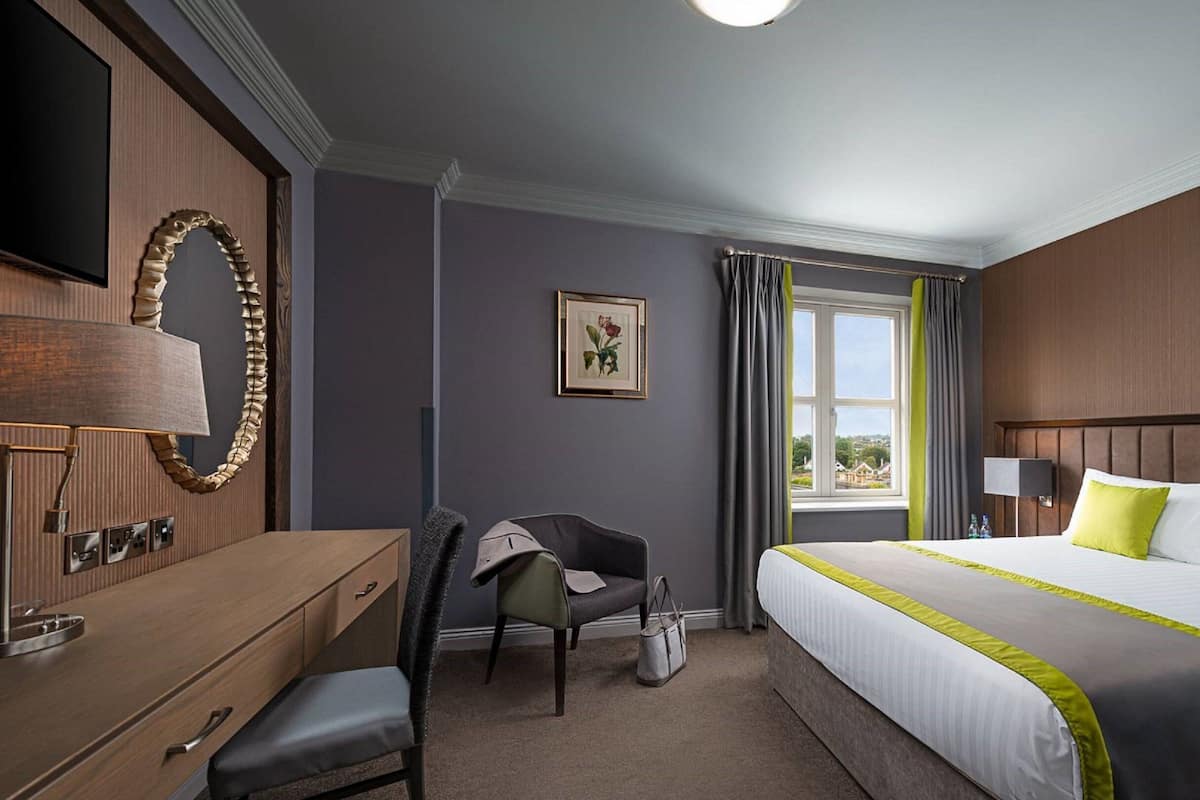 Een hotelkamer met een bed, een bureau, een stoel, een dressoir, een spiegel, een raam met gordijnen en een lijst met een afbeelding aan de muur.