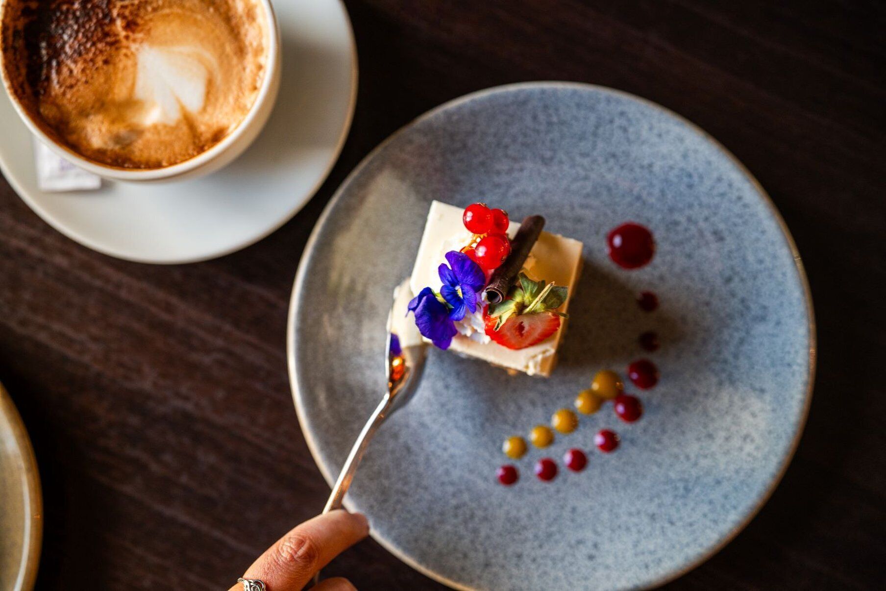 Un postre en un plato con una cuchara, una taza de café y una mesa de madera oscura.